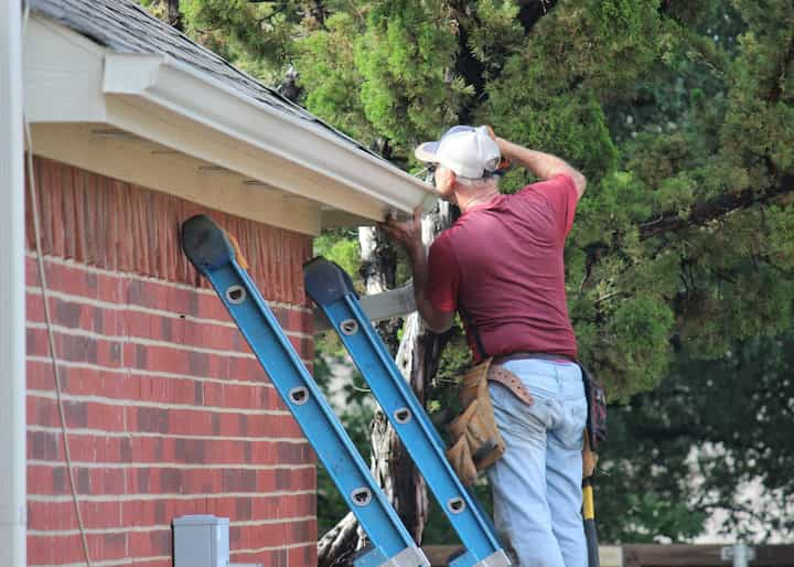 A professional emergency roof repair contractor wears grey hat on ladder in Whittier, California